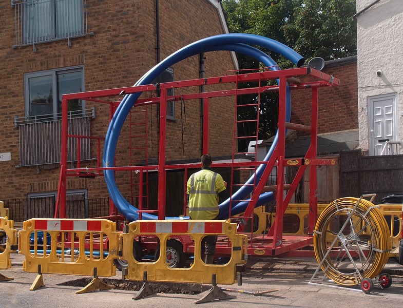 Worker in yellow vest stands next to a large blue pipe coil on a street.