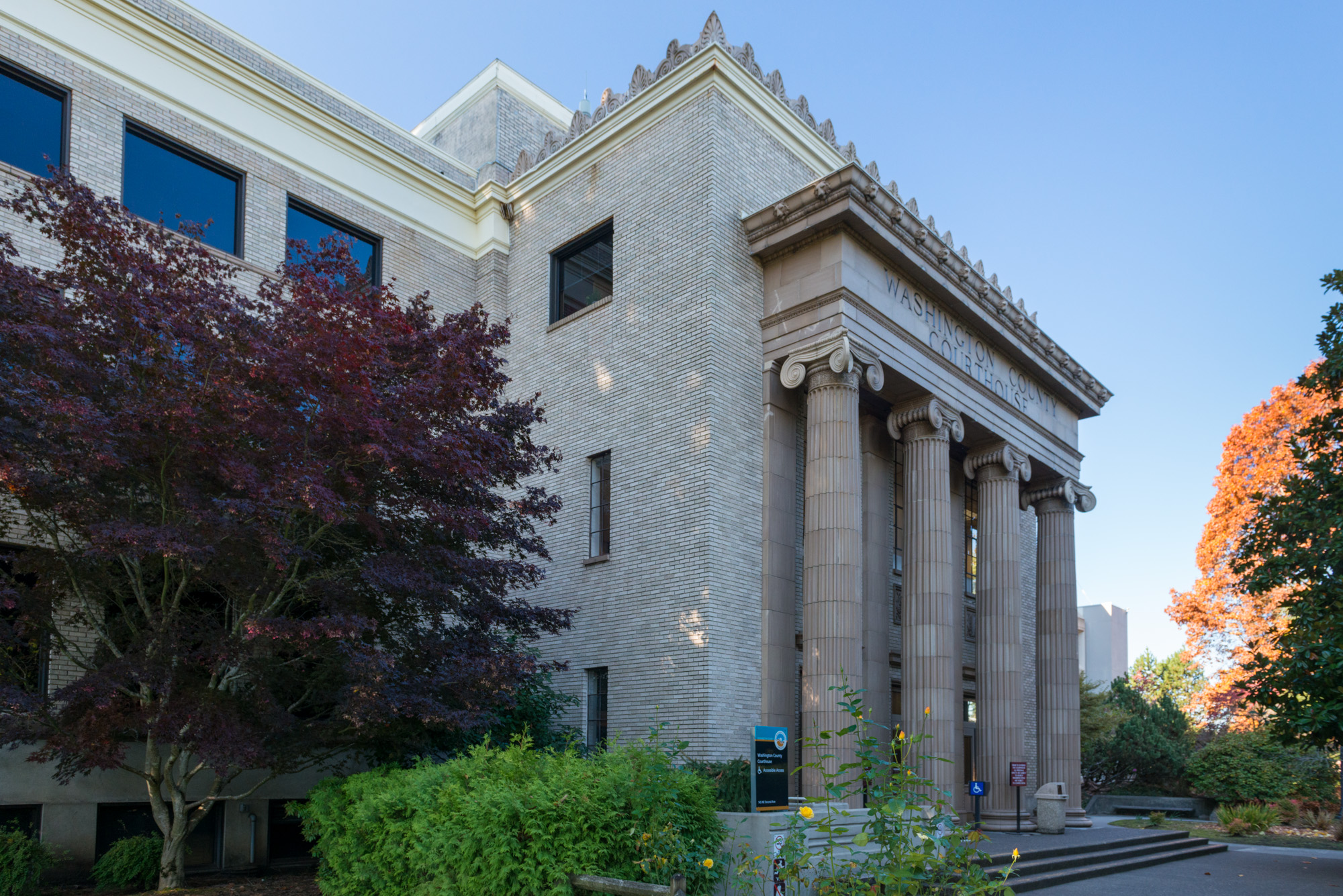 Neoclassical courthouse with tall columns and a red-leafed maple tree against a blue sky.