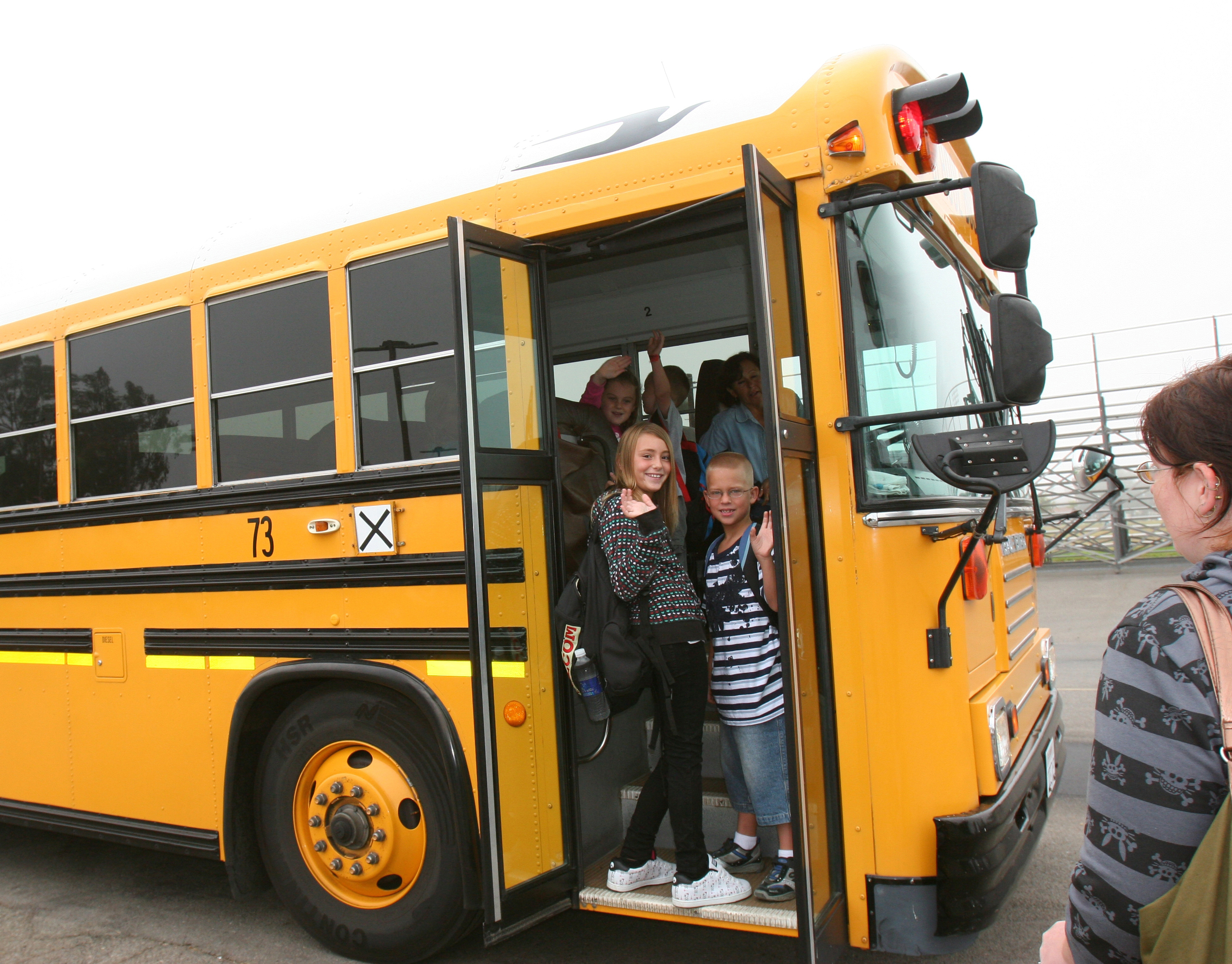Two young children wave from the open doorway of a yellow school bus.