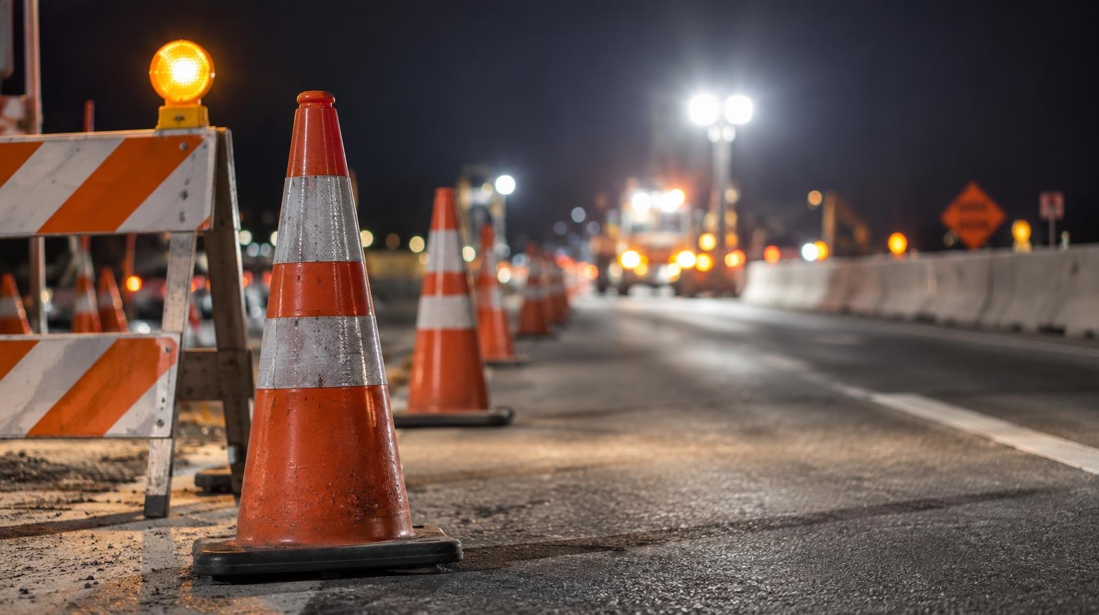 Orange traffic cones and barricades lining a closed highway lane at night under bright work-zone lights.