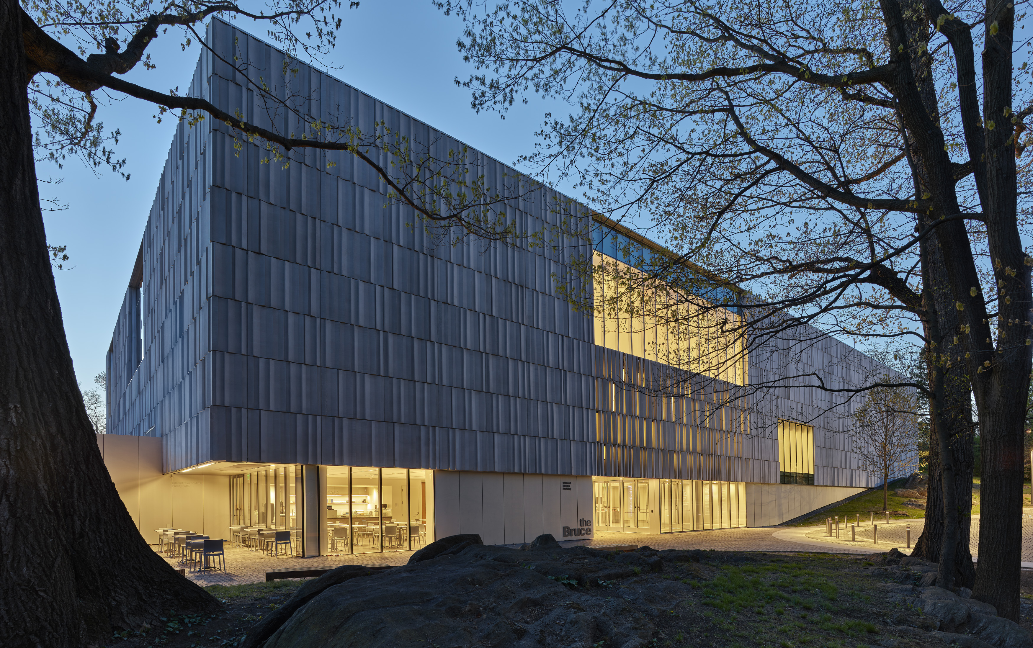 Modern museum building with metallic paneled facade illuminated at dusk, framed by mature trees