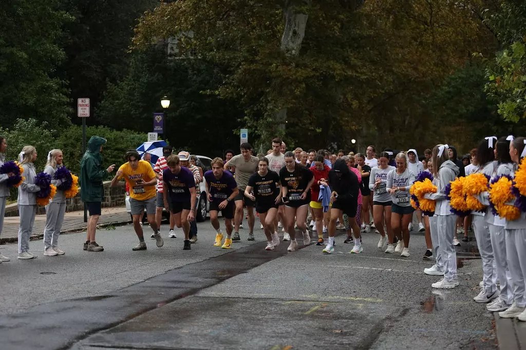Runners at a race start line on a street, flanked by cheerleaders with gold and purple pom-poms.
