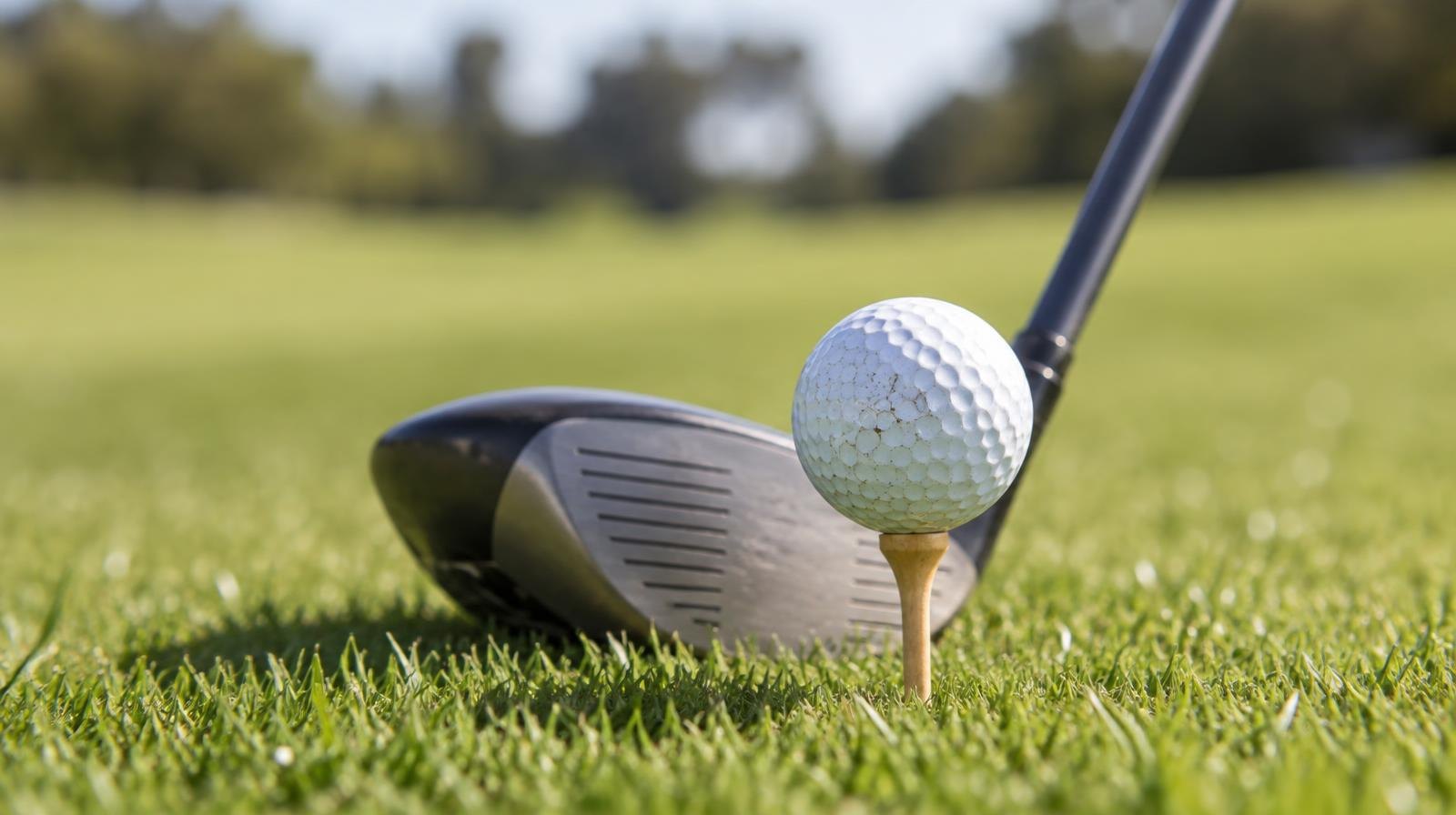 A golf ball on a wooden tee in short grass, with a club head positioned behind it.