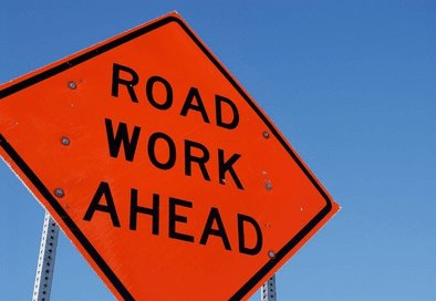 An orange diamond-shaped Road Work Ahead construction warning sign against a blue sky.