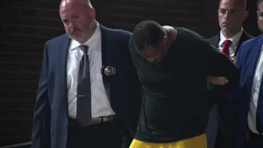 A man in a green shirt is escorted by two suited law enforcement officers outdoors at night.
