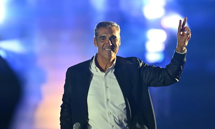 A man in a dark blazer raises two fingers in a victory sign against blue stage lighting.