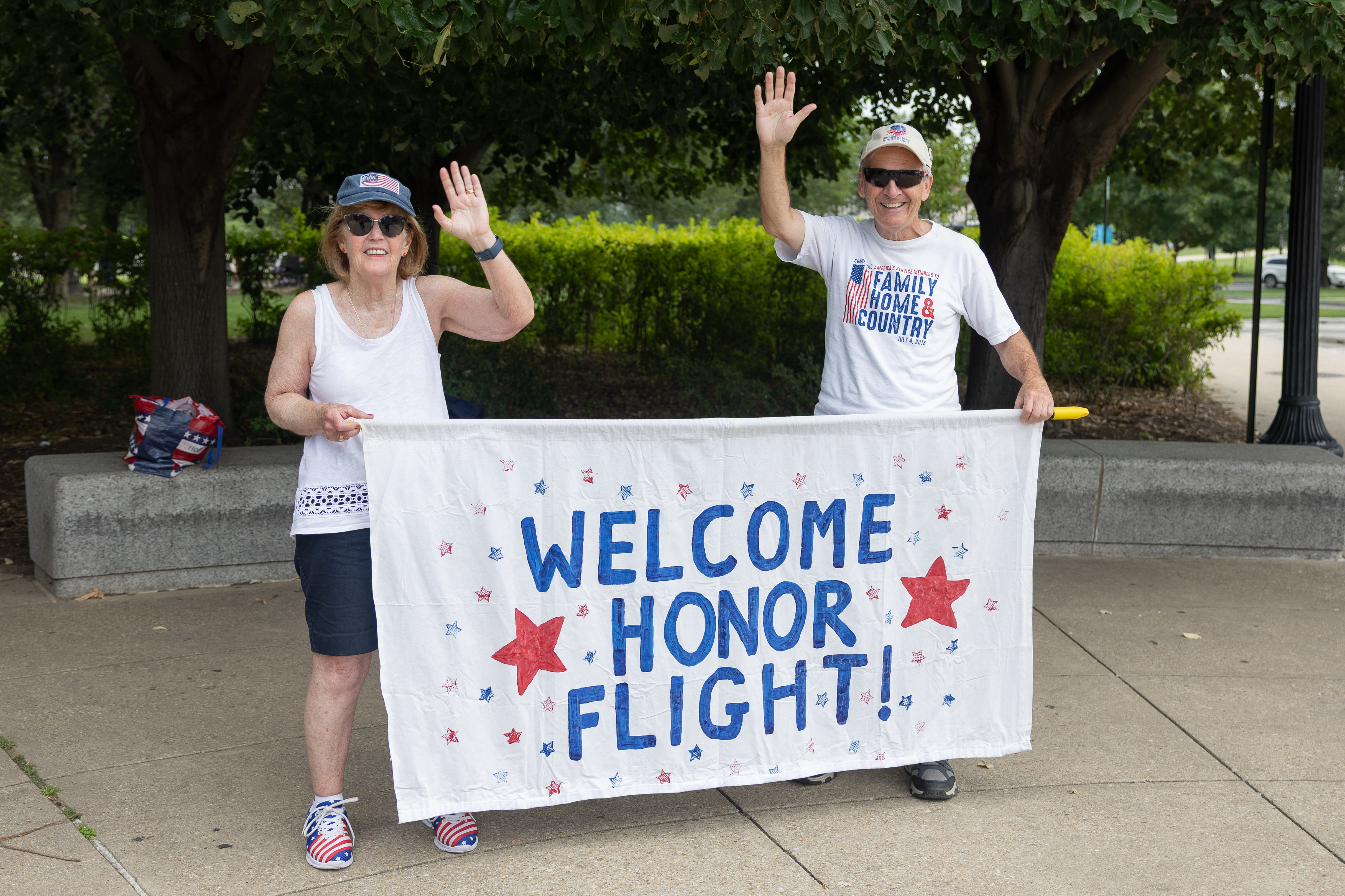 Two people wave and hold a hand-painted 'Welcome Honor Flight!' banner with red and blue stars.