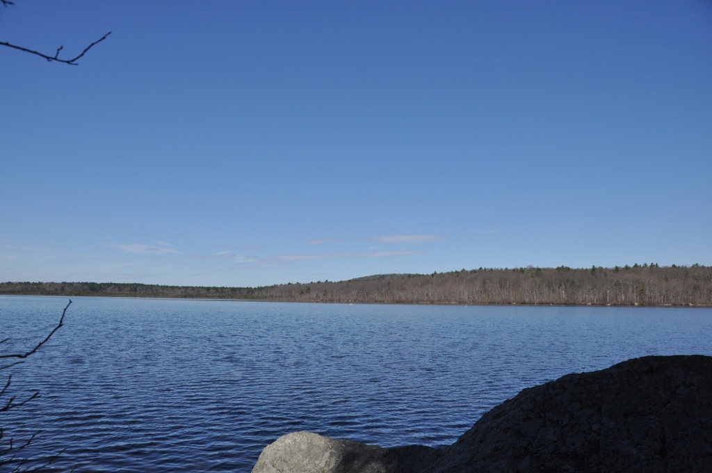 Calm blue pond with rocky shoreline and forested hill in the distance.