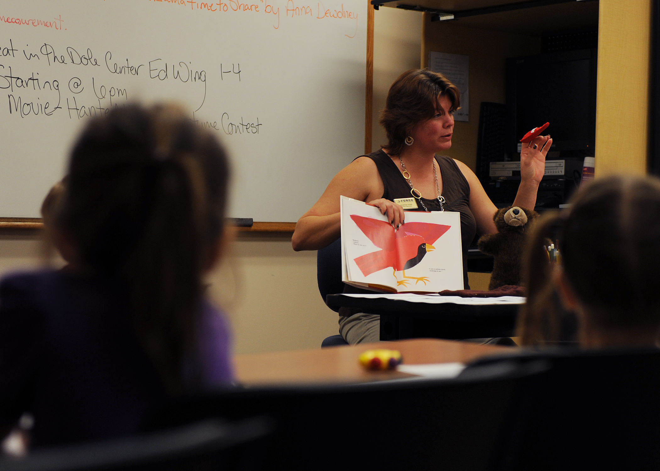 A woman holds up a colorful picture book while reading to young children at desks.