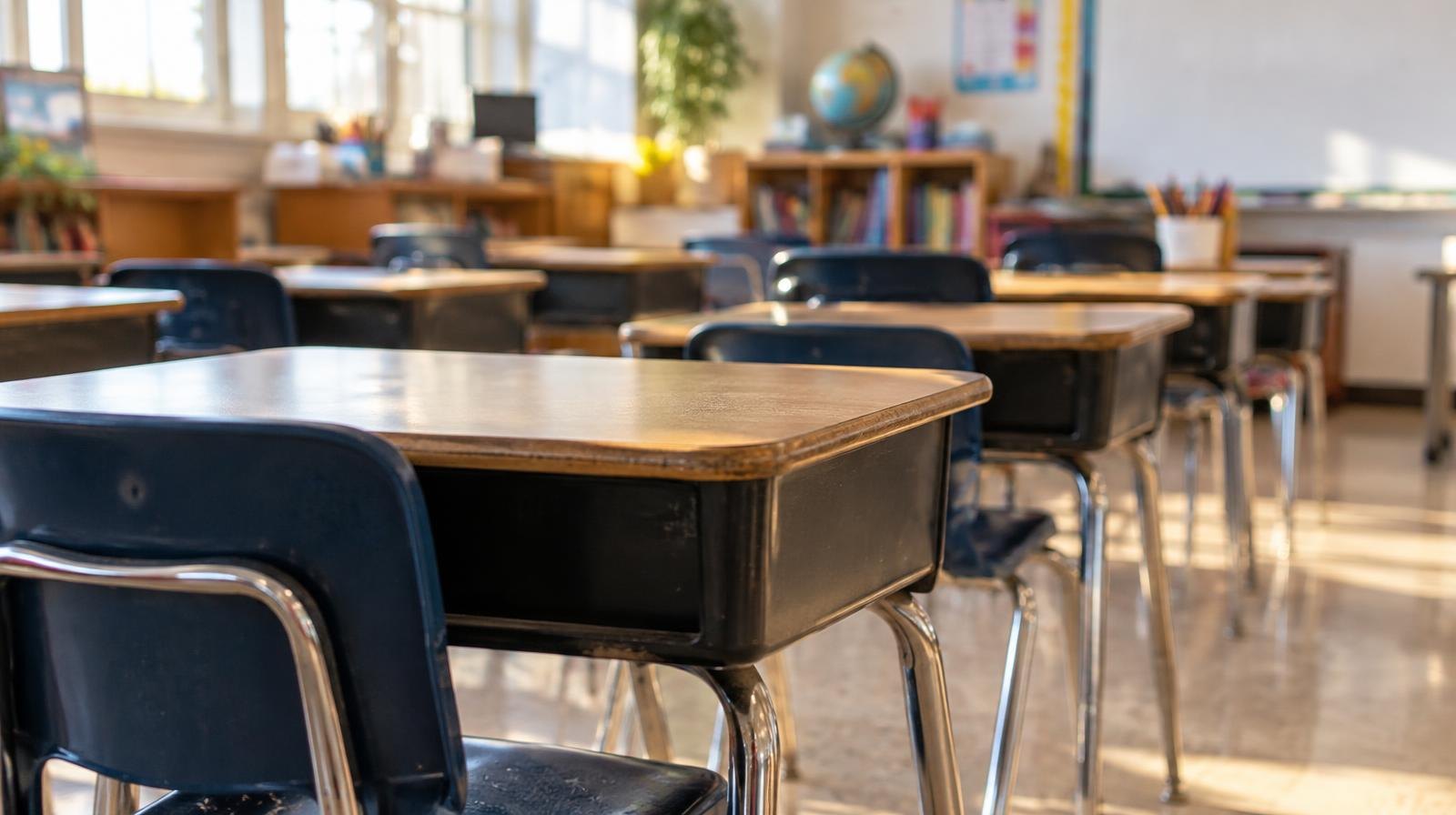 Rows of empty student desks in a bright, sunlit classroom.