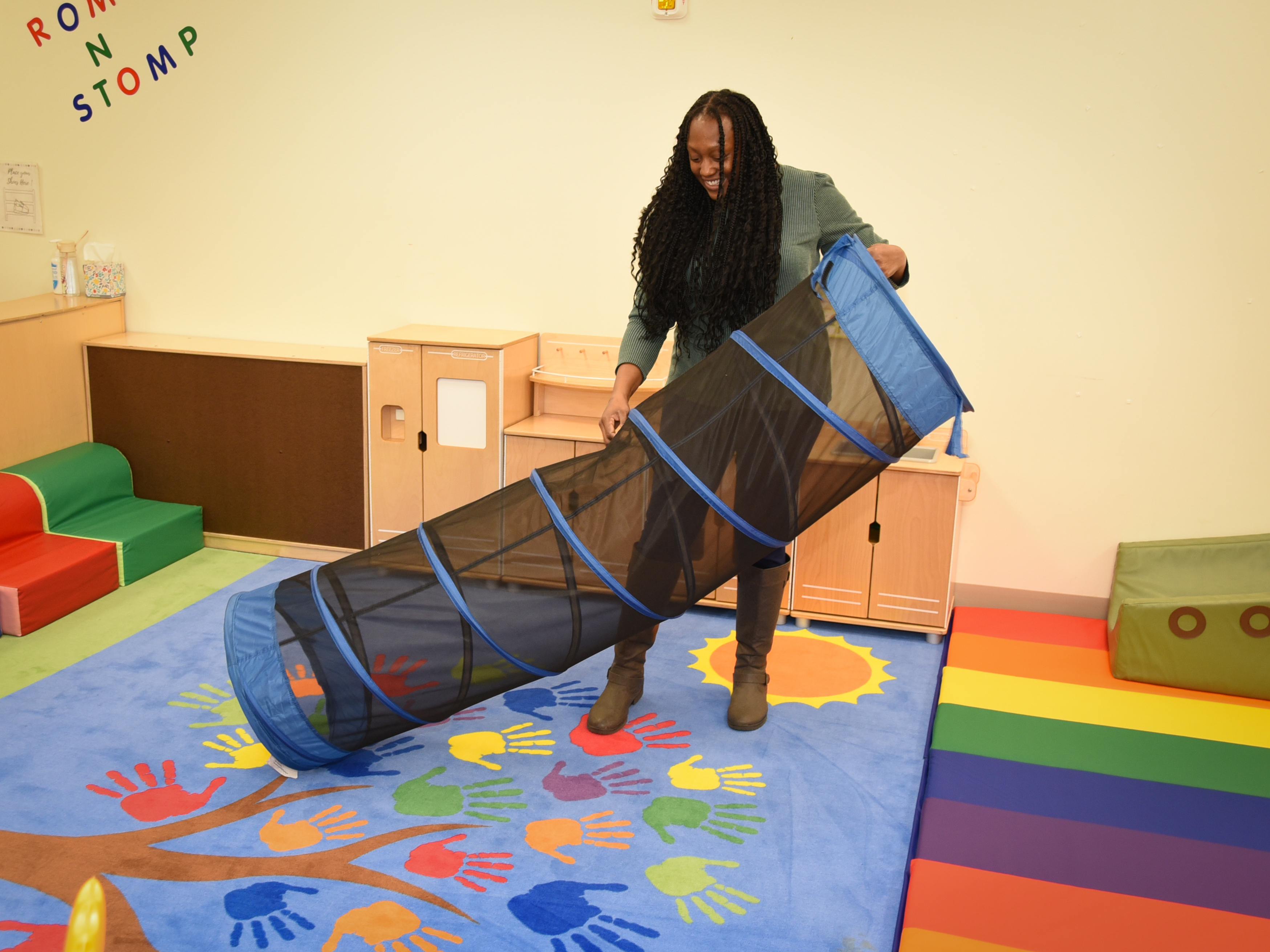 A woman sets up a mesh play tunnel on a colorful rug in a children's play room.