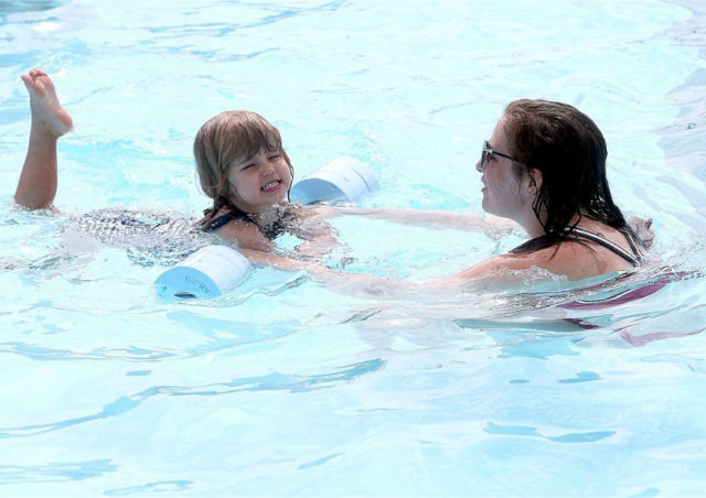 A young child using a pool noodle flotation device while an adult assists in an outdoor swimming pool.
