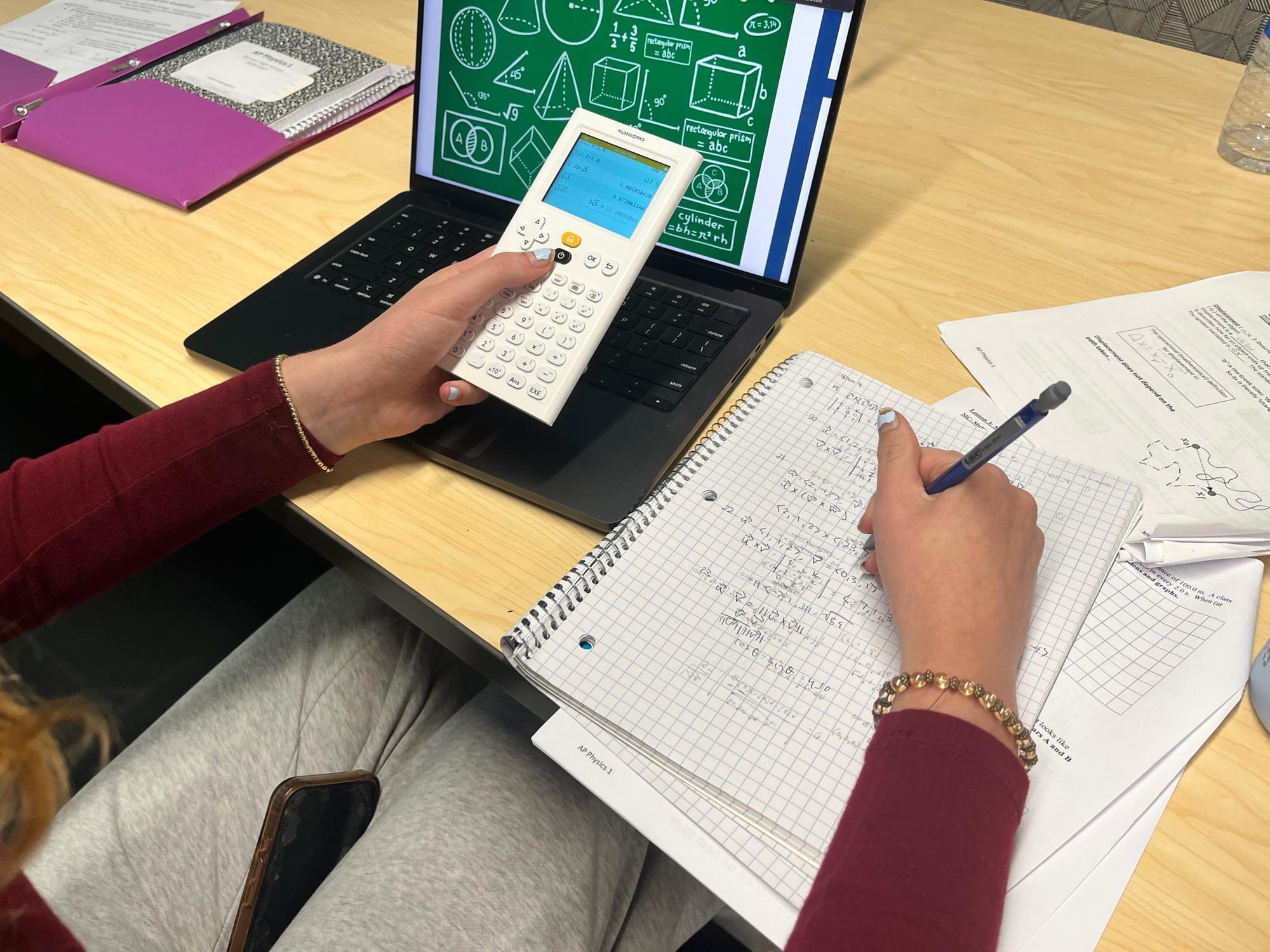 Hands holding a calculator and pen over a notebook with math equations on a school desk.