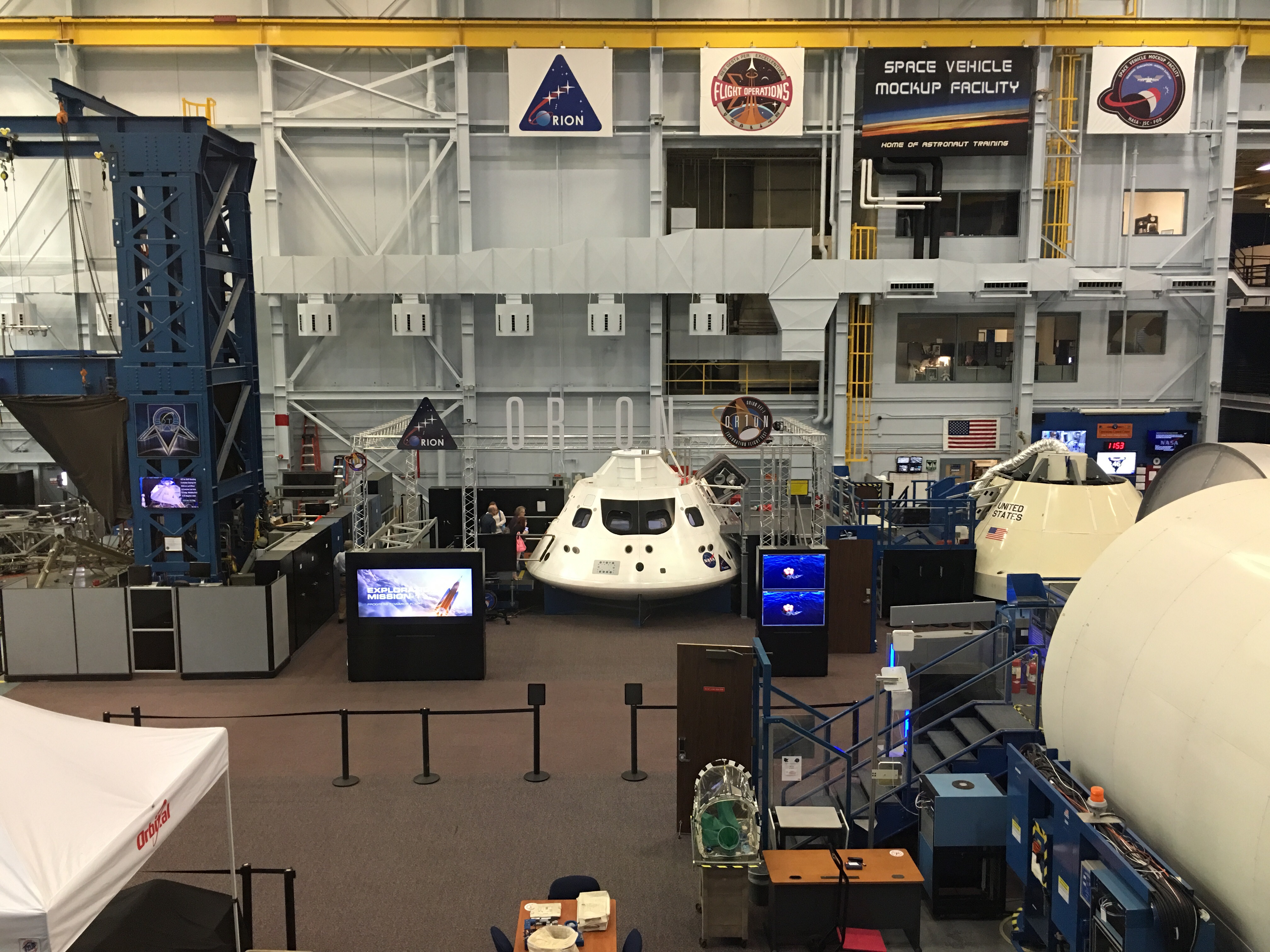 Orion spacecraft capsule mockup inside a large NASA facility with mission banners overhead.