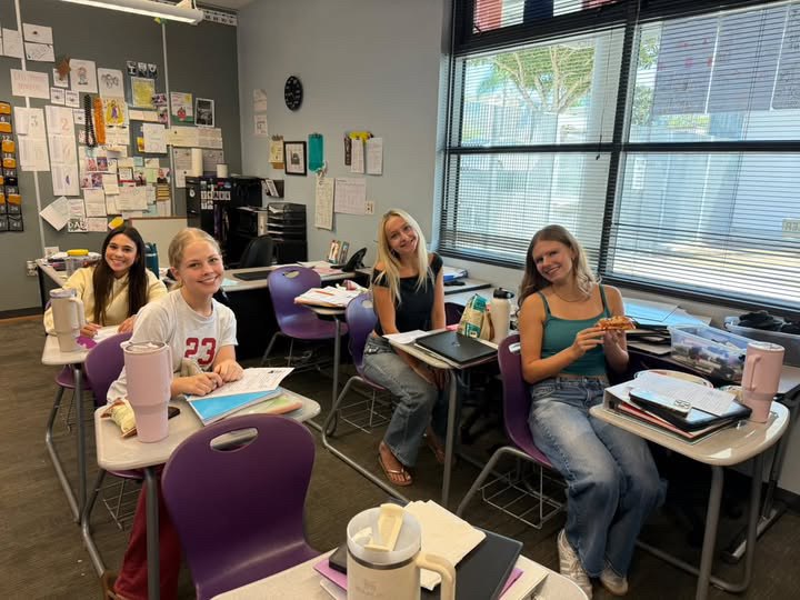 Four students seated at purple desks in a classroom, smiling at the camera.