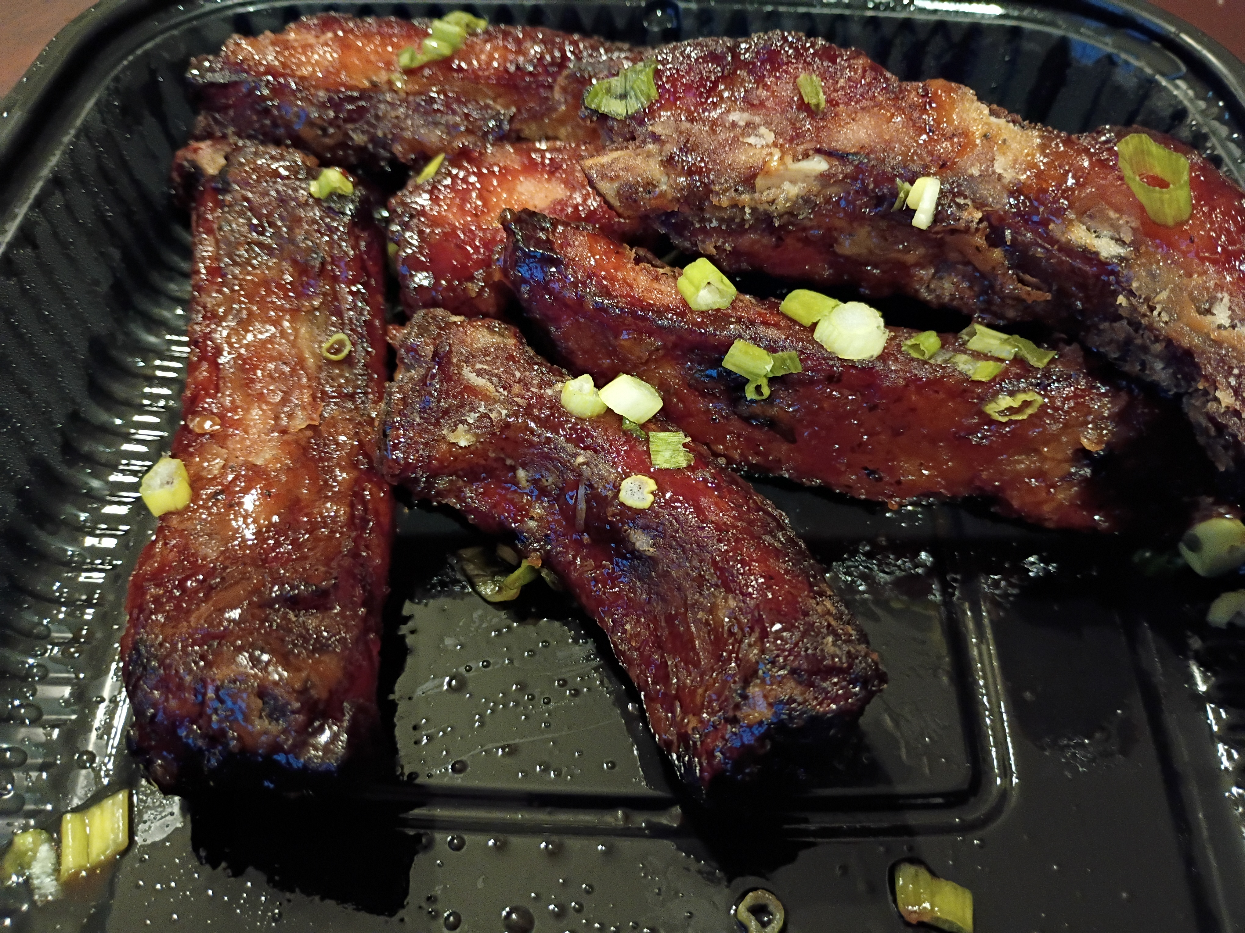 Glazed barbecue ribs in a black takeout tray, garnished with green onions.