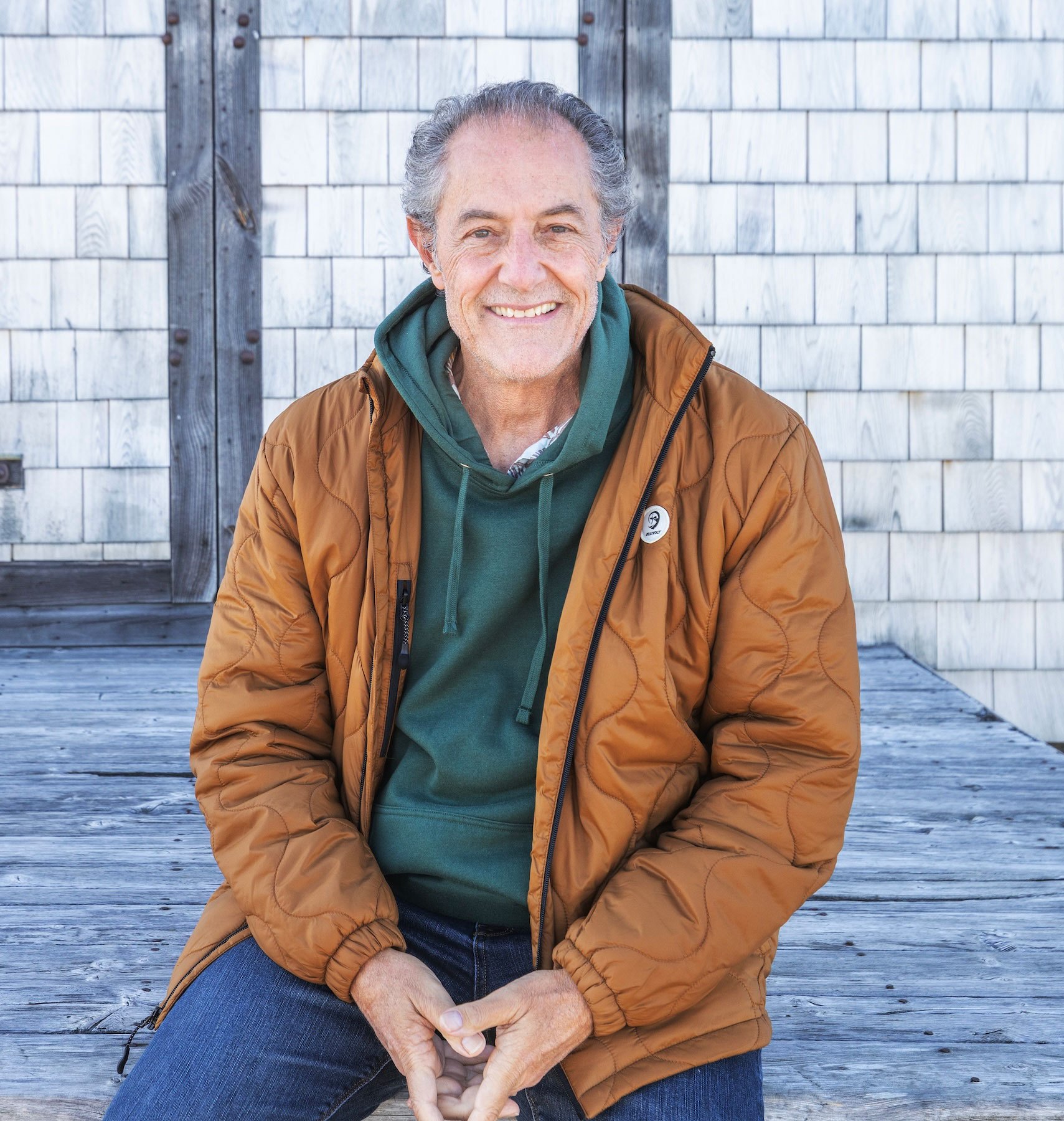 A smiling older man with gray hair sits on a wooden deck wearing a tan quilted jacket over a green hoodie.