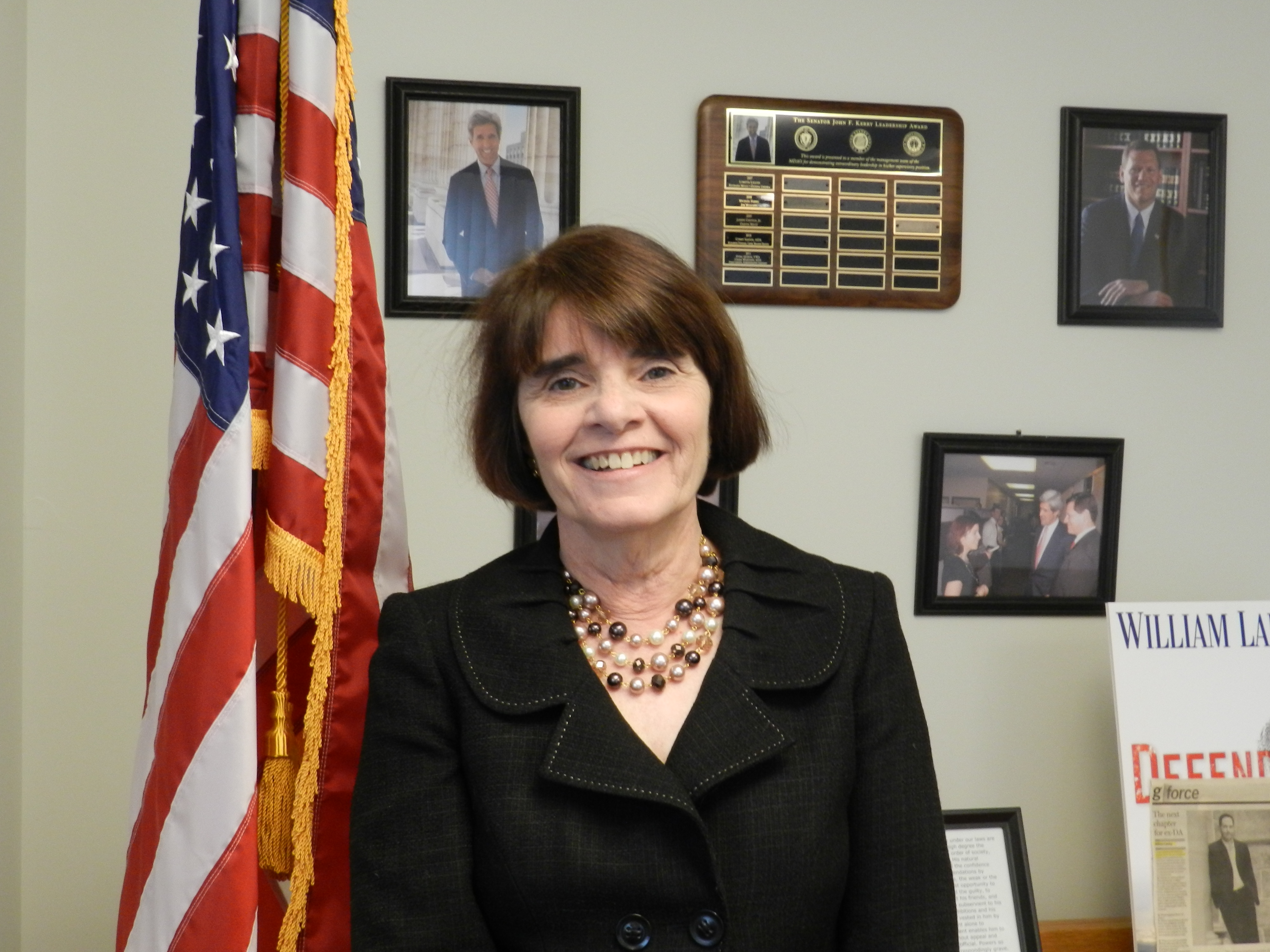Woman in dark blazer stands before an American flag in an office.