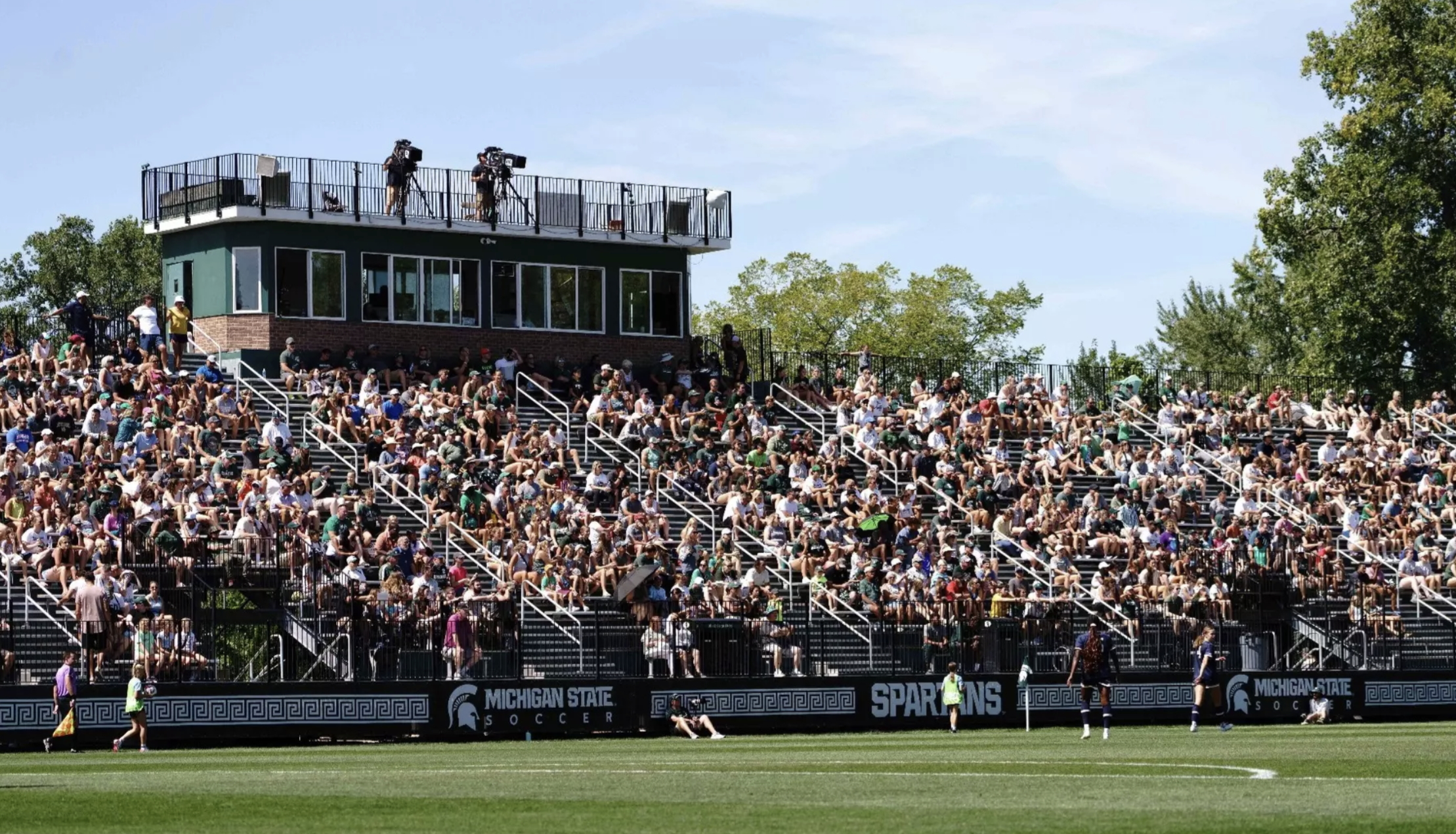 A packed crowd fills the bleachers at DeMartin Stadium, with a press box overlooking the stands and players warming up on the field below.