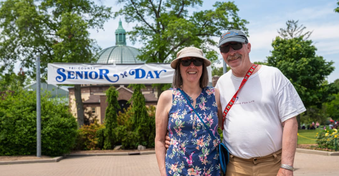 A smiling man and woman pose together in front of a "Tri-County Senior Day" banner at the Detroit Zoo, with the zoo's domed pavilion visible in the background.