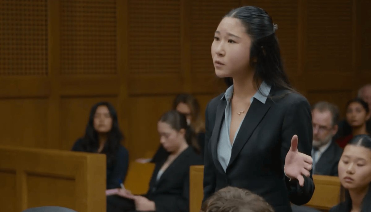 Young woman in a dark blazer stands and speaks in a wood-paneled courtroom-style room.