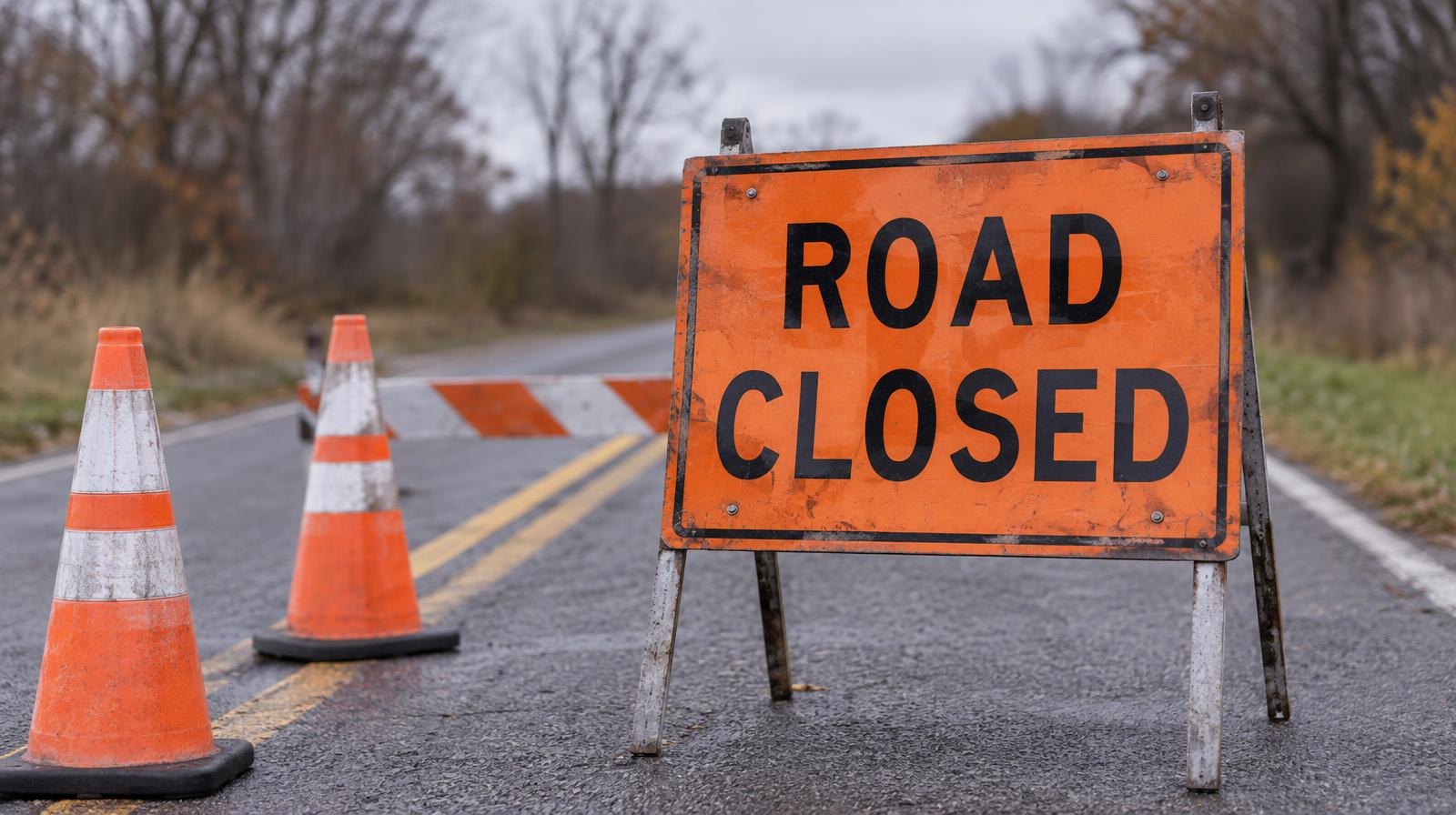 Orange road closed sign and traffic cones blocking a two-lane rural road on an overcast day.