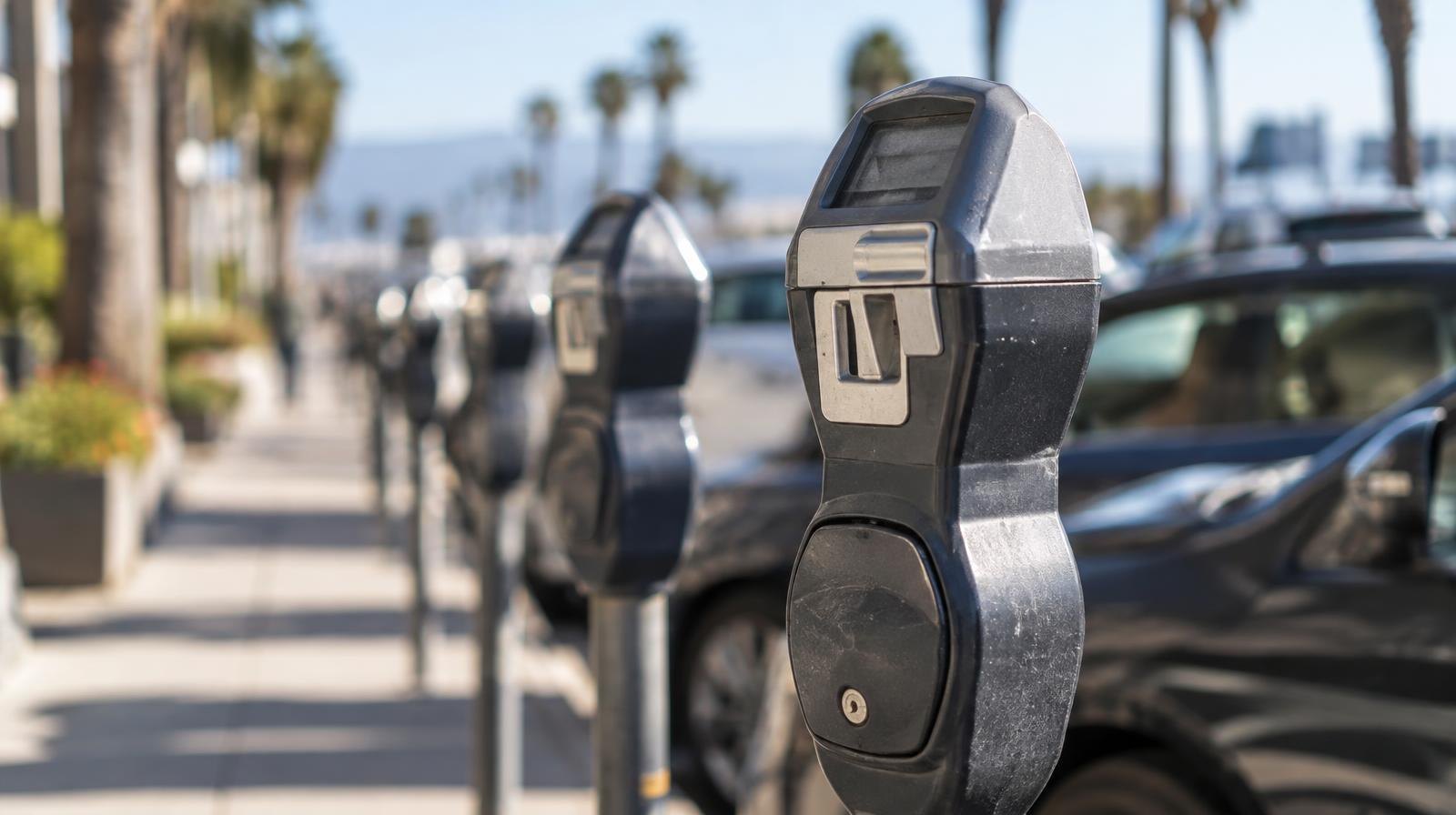 Modern parking meters lined up on a sunny city sidewalk beside parked cars.