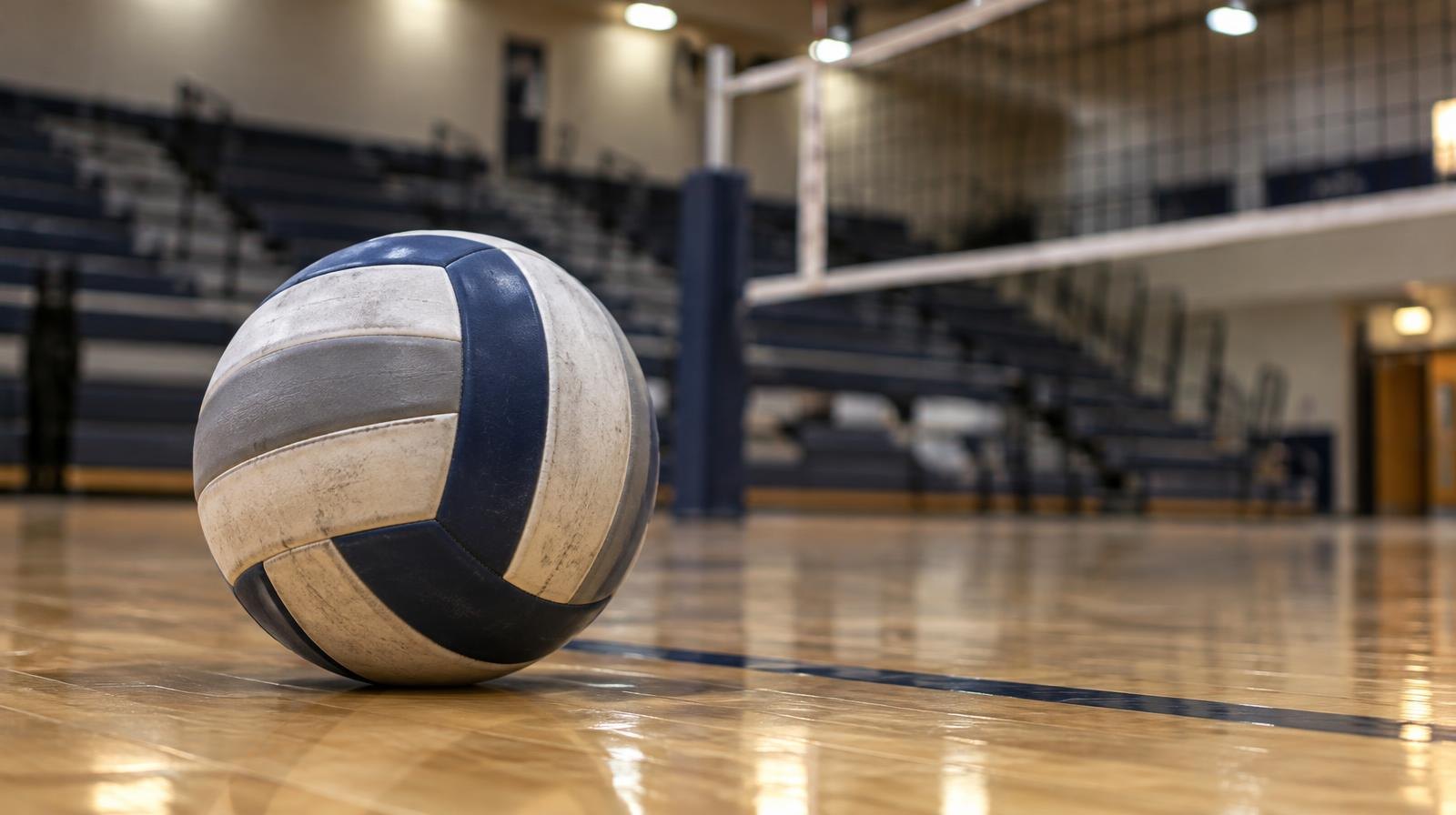 A volleyball resting on a hardwood gym floor near the net, bleachers blurred in the background.