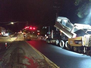 Nighttime road paving with a dump truck, paving machine, and orange traffic cones.