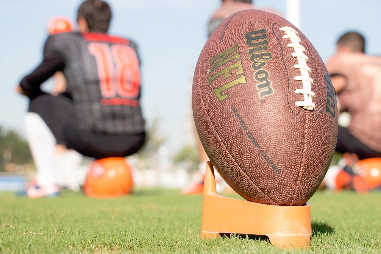 A football rests on an orange kicking tee on a grass field, with players and orange helmets blurred in the background.