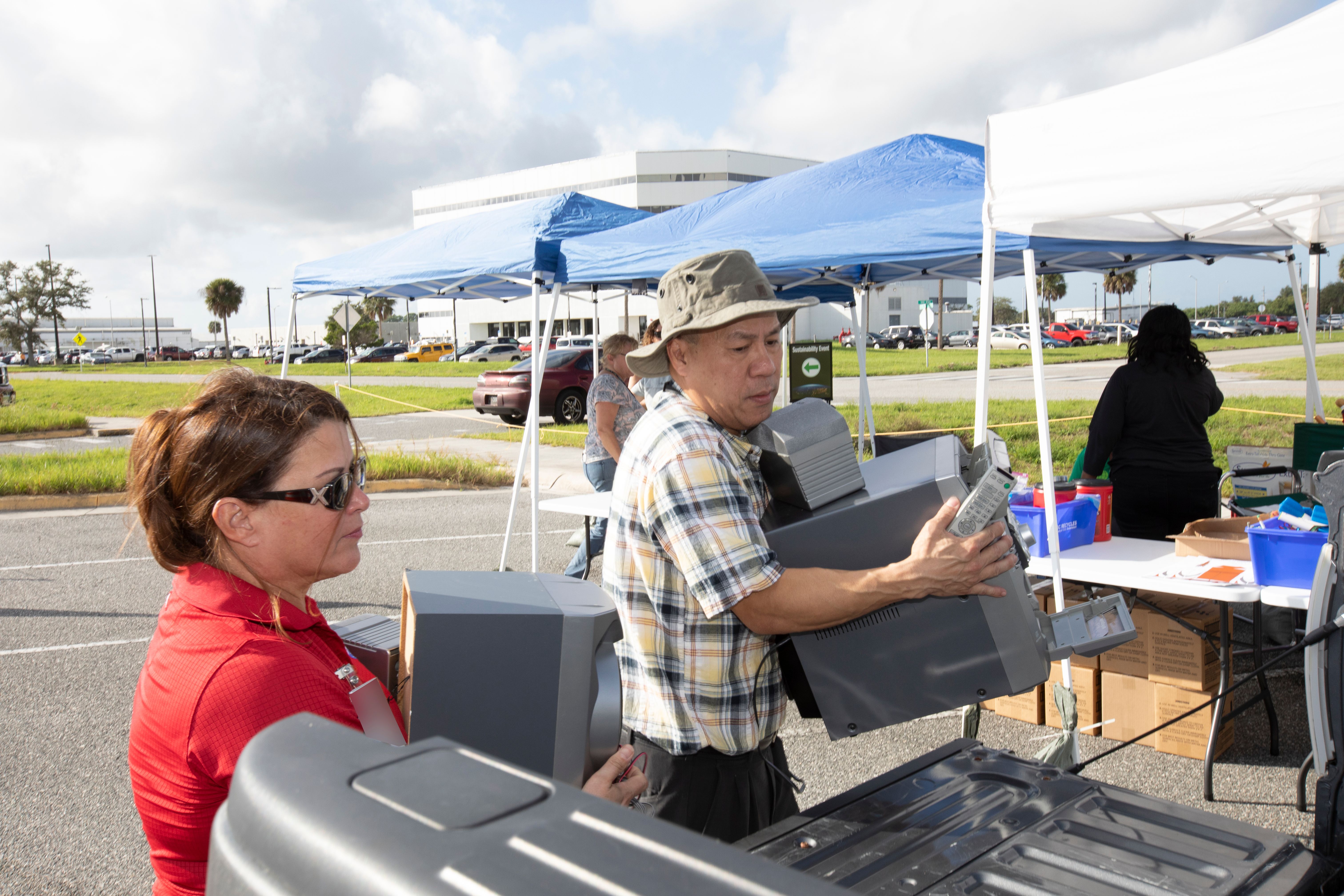 Two people hand off grey trays at an outdoor recycling drop-off event under blue canopy tents.