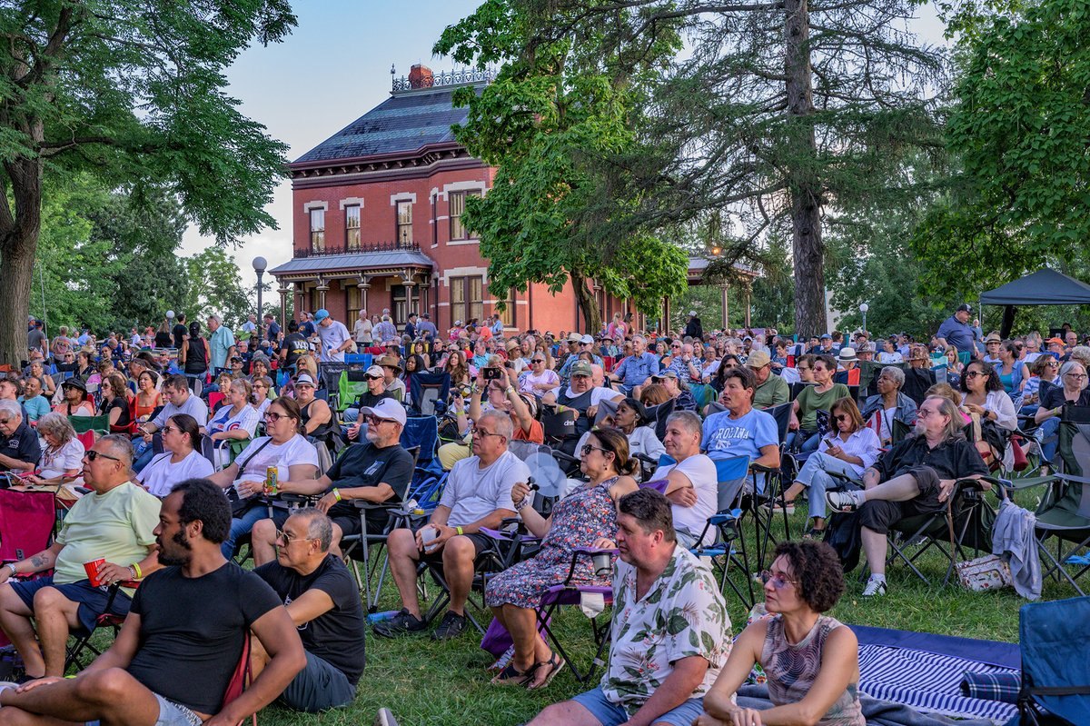 Large crowd seated in lawn chairs on a grassy field in front of a historic red brick building during an outdoor evening event.