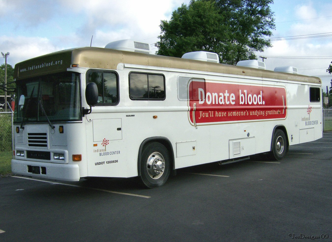 Large white mobile blood donation bus with a red banner parked outdoors.