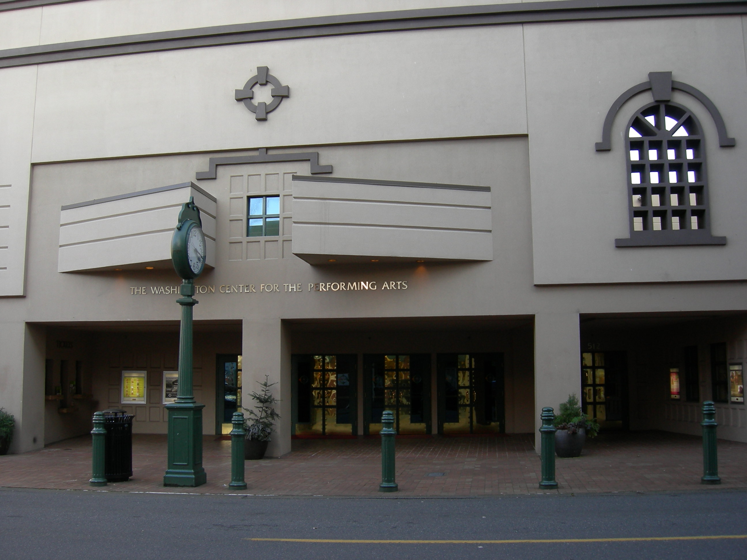 Entrance of a performing arts center with glass doors, green bollards, and a street clock.