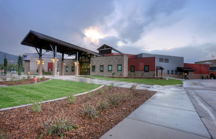 Modern school building exterior with covered entrance canopy, stone facade, and mountains in the background.