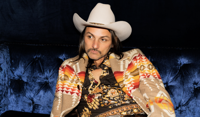 Duane Betts, wearing a white cowboy hat and a Southwestern-patterned jacket, sits on a blue velvet couch.