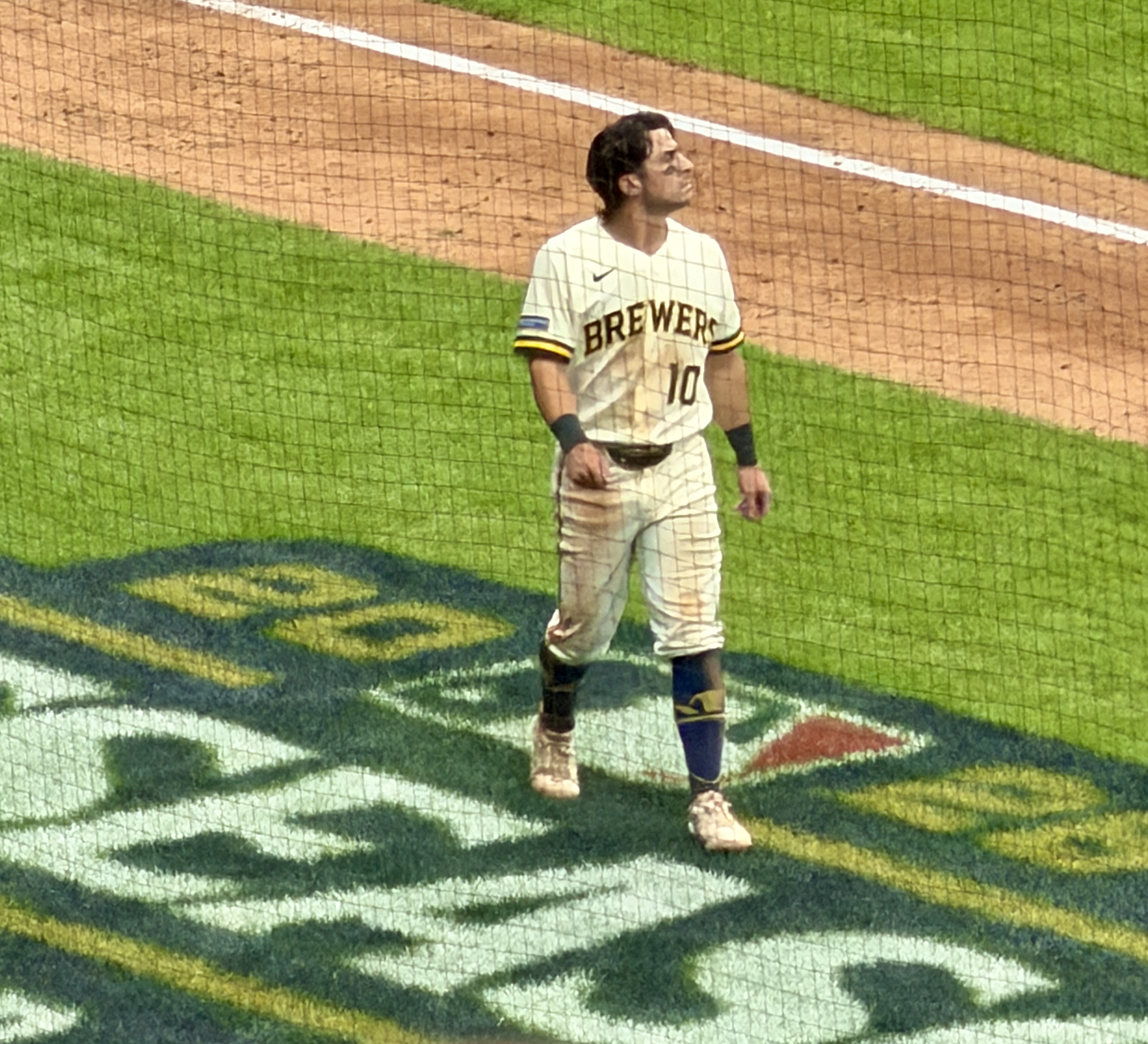 Baseball player in cream Brewers jersey number 10 standing on a field near the dugout.