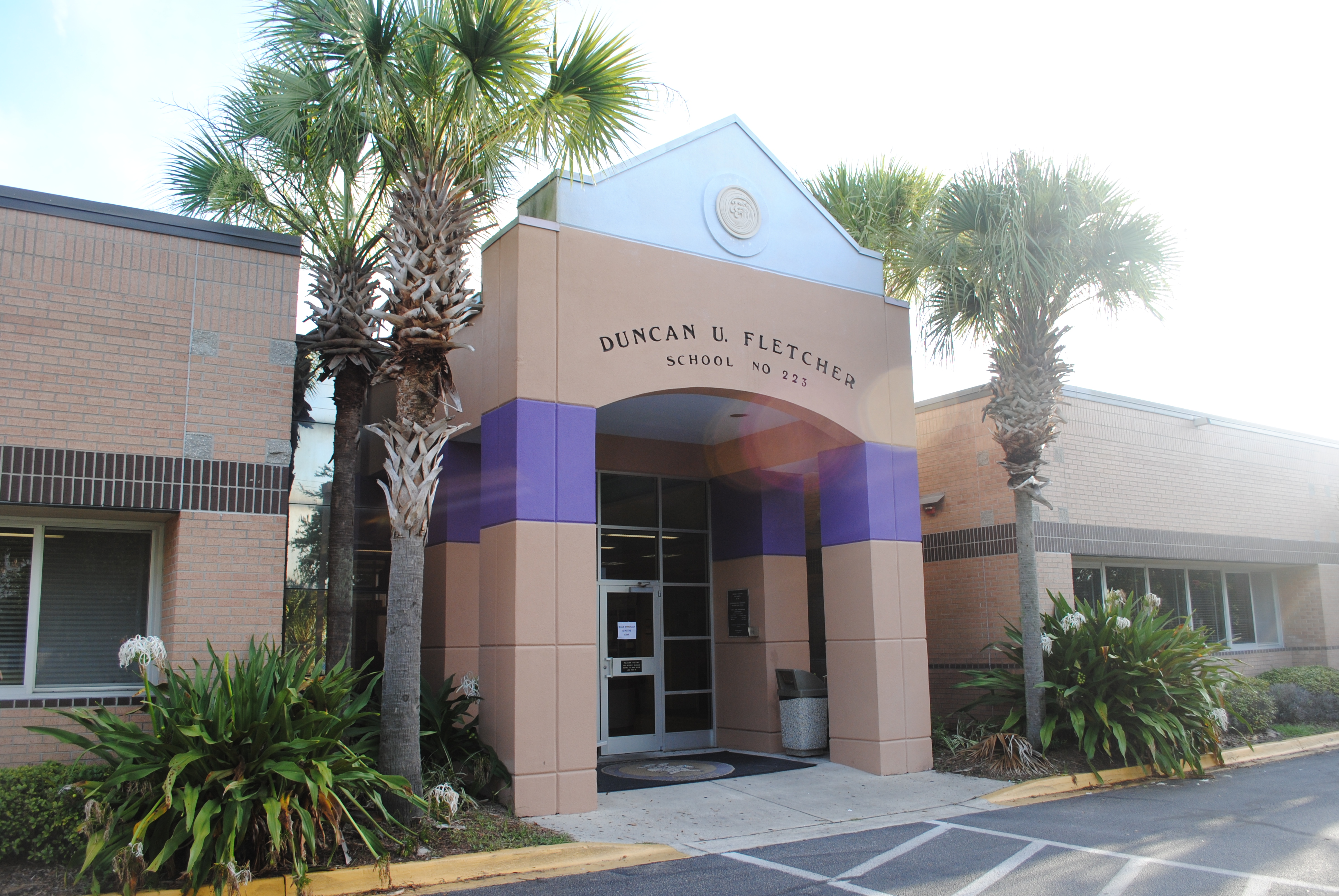 School entrance with tan and purple brick facade, palm trees, and a covered portico.