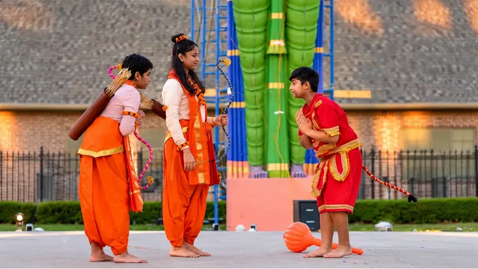 Three children in traditional Indian costumes performing outdoors with a large colorful effigy in the background.