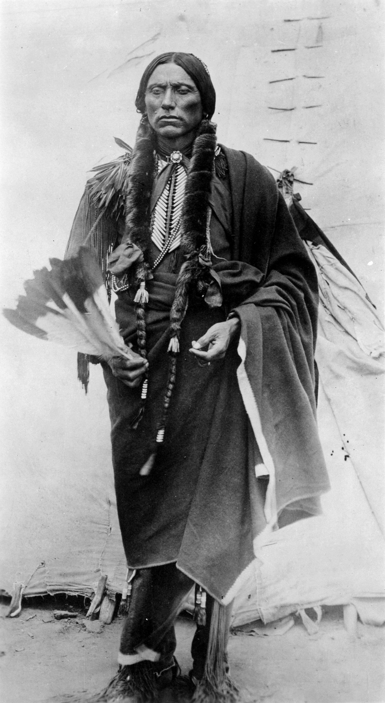 Black-and-white portrait of a Native American man in traditional dress standing before a tipi.