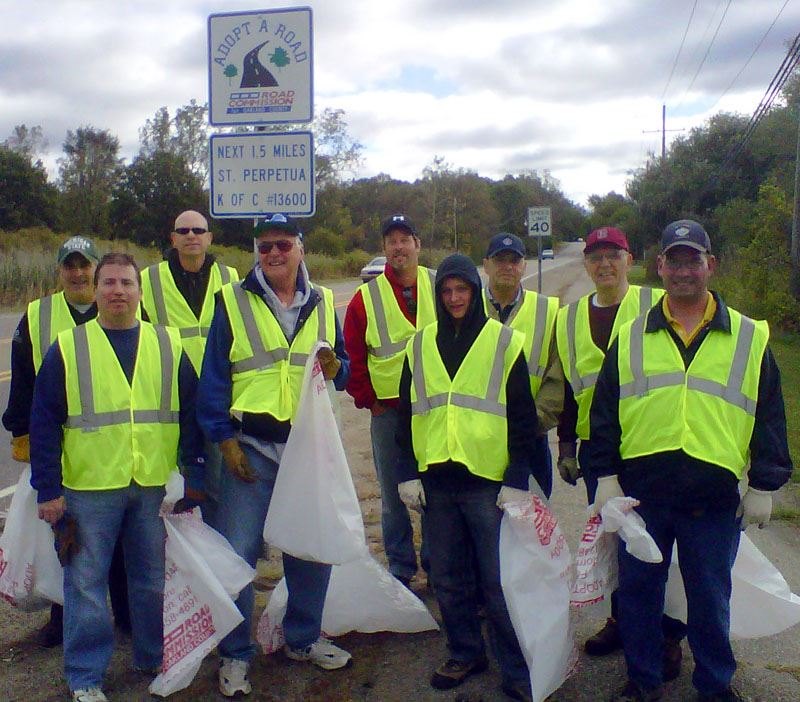 Volunteers in yellow vests holding white trash bags stand beside an Adopt-A-Road sign.