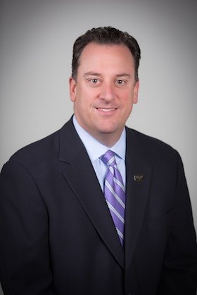 Professional headshot of a man in a dark suit and striped tie against a gray background.