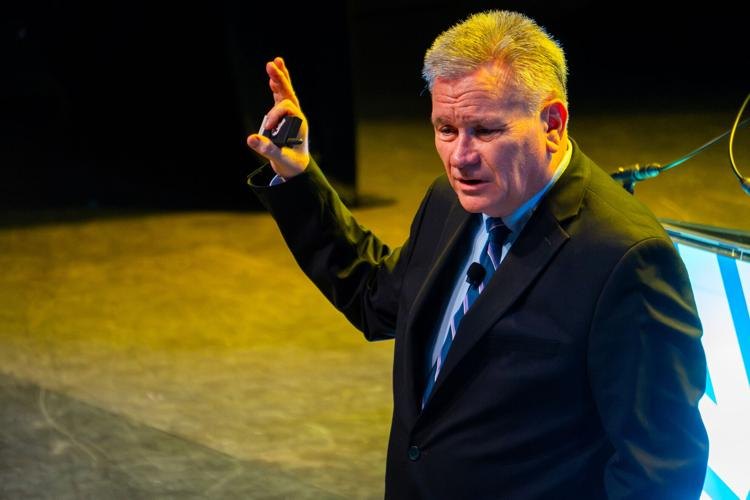 A man in a dark suit gestures while speaking at an indoor event stage.