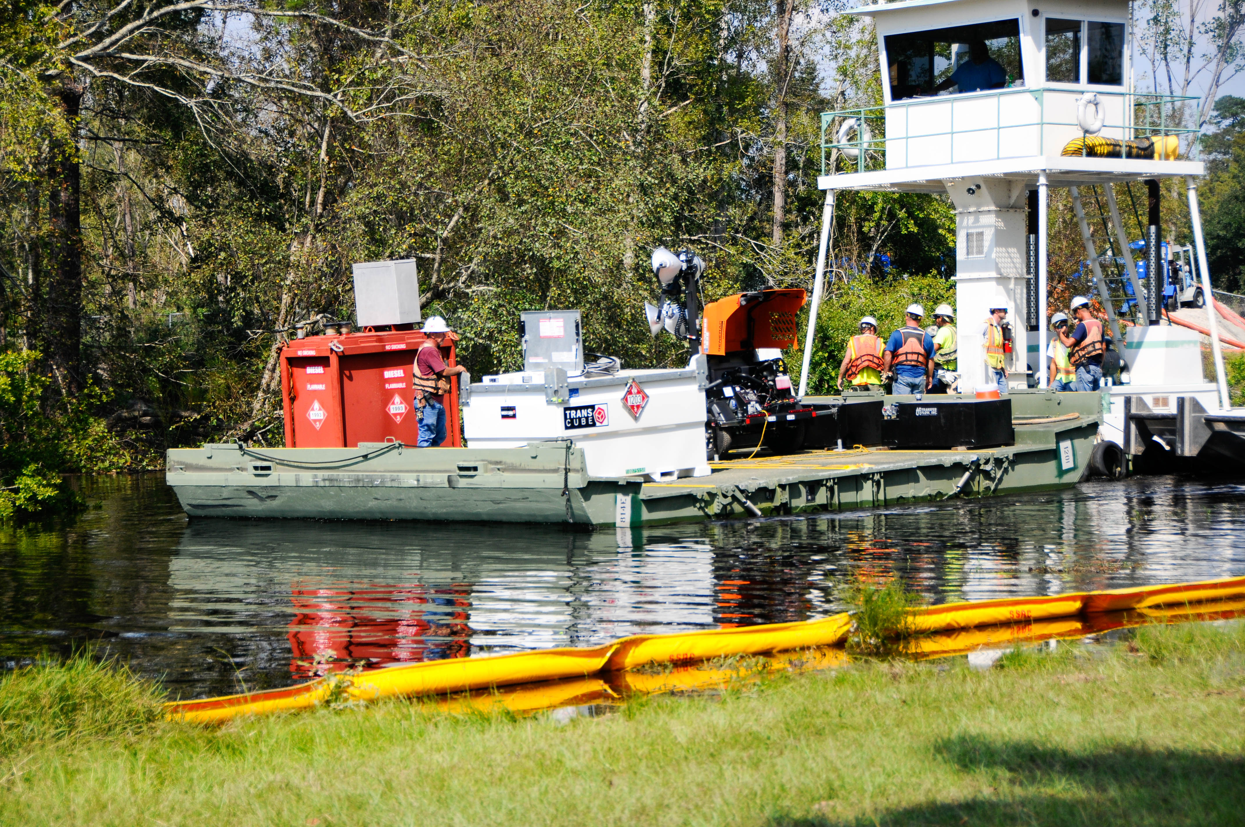 Workers in safety vests and hard hats aboard a flat-bottomed boat on a waterway.