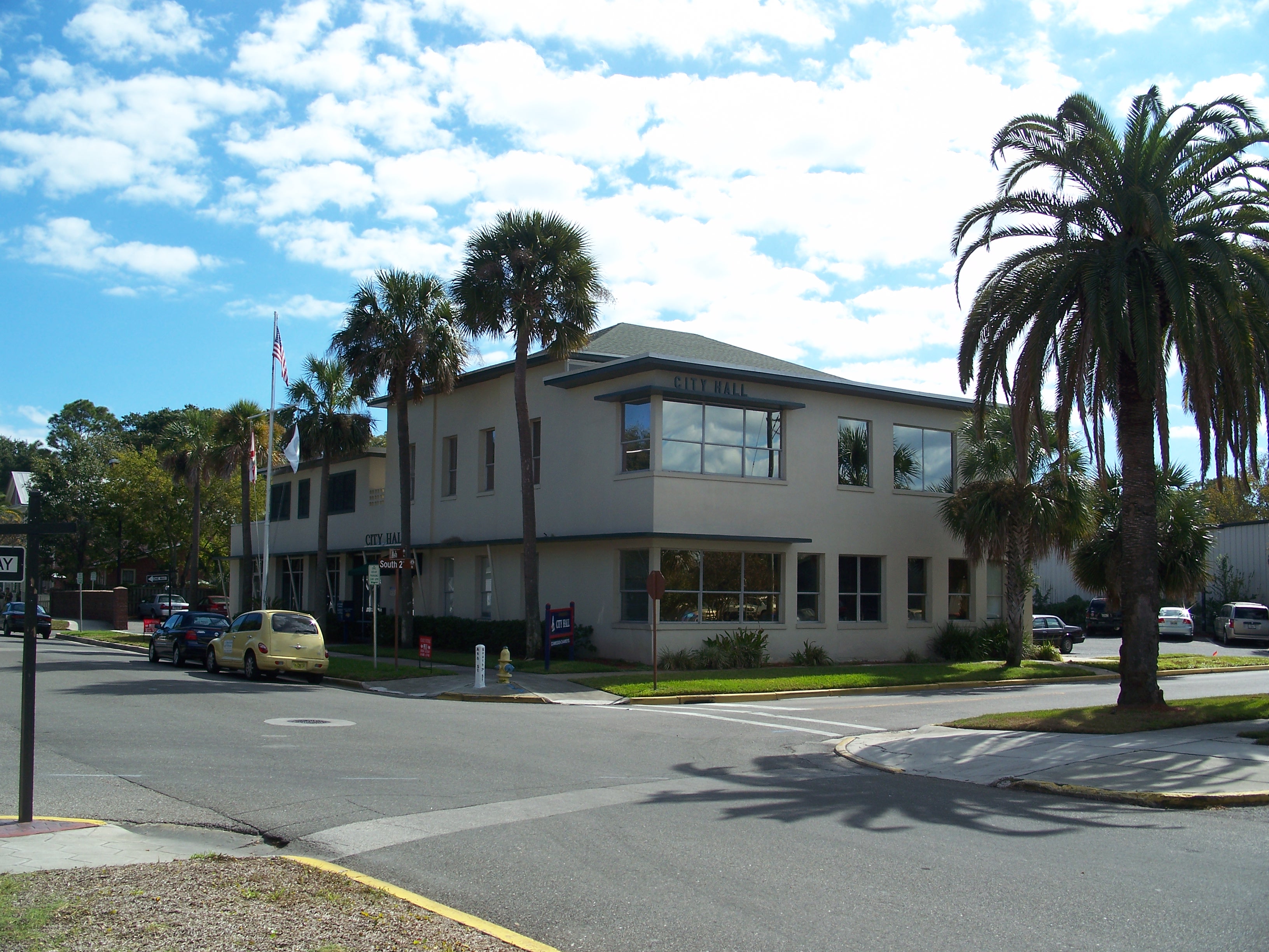 Two-story tan municipal building at a street corner with palm trees and a flagpole.