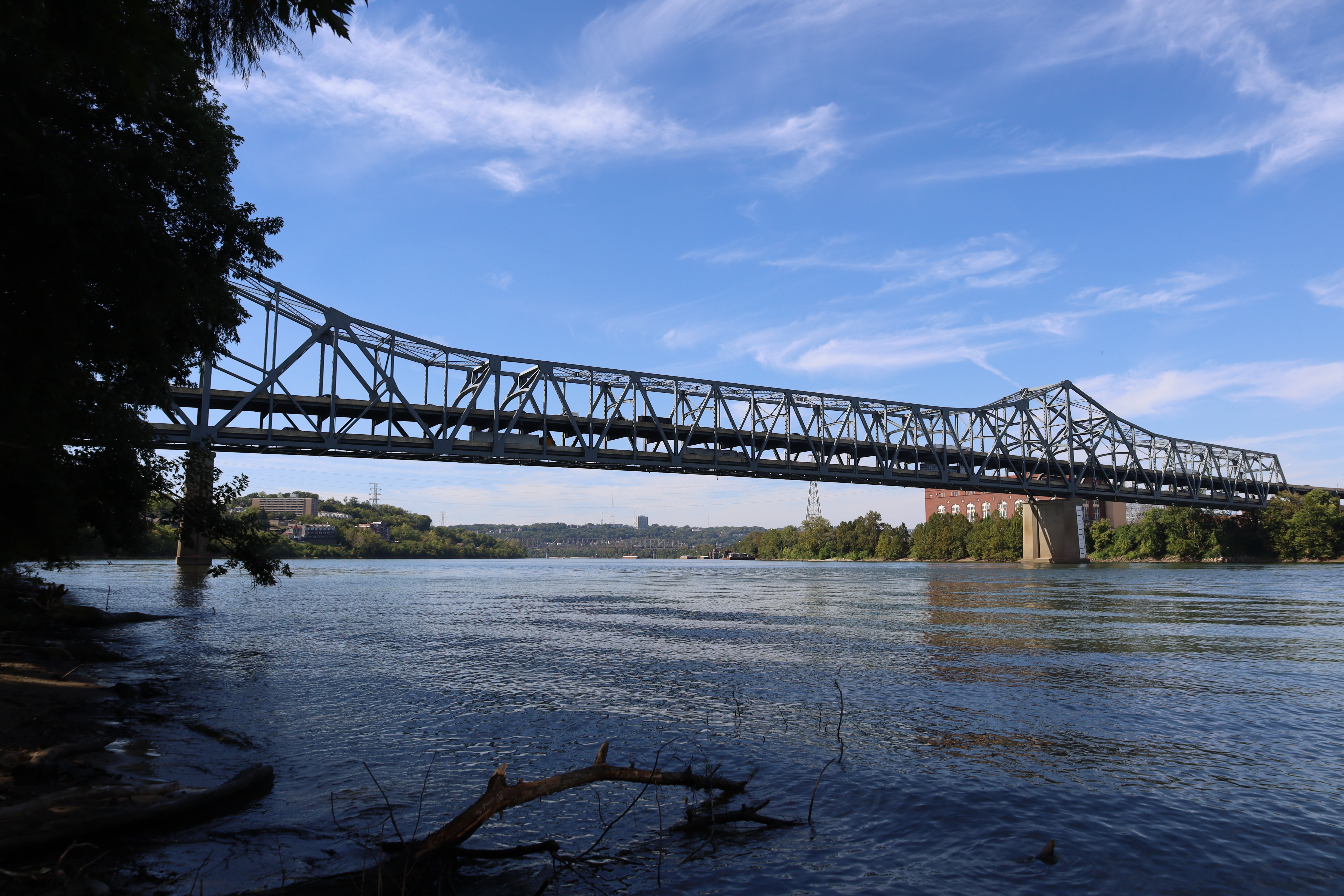 Steel truss bridge over a wide river viewed from a low riverbank under a blue sky.