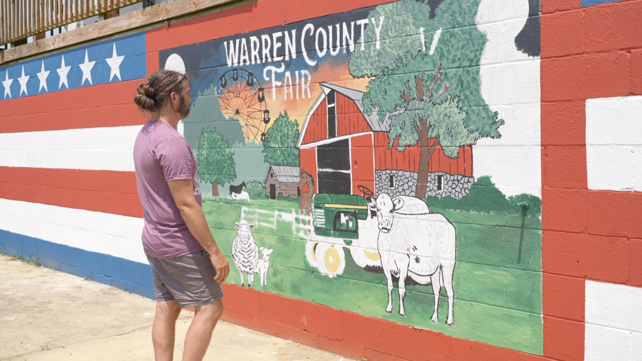 Person in purple shirt stands before a large painted fair mural on a brick wall.