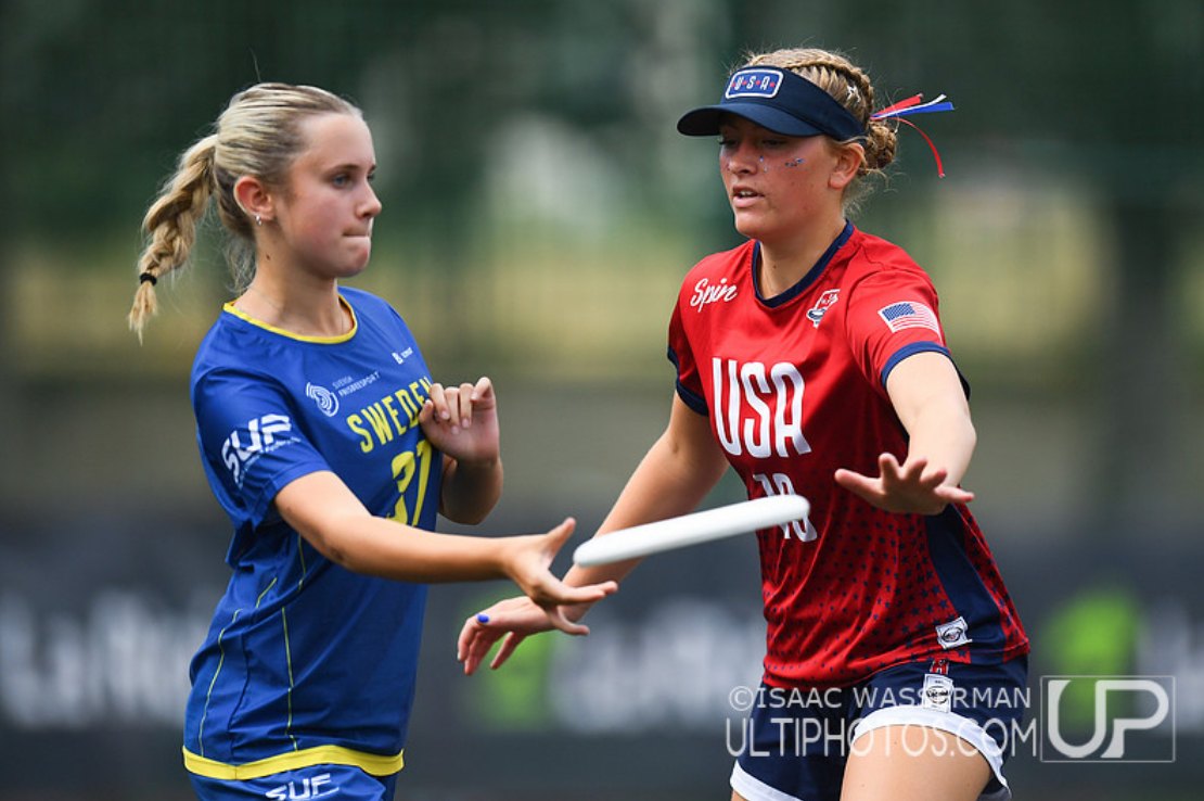 Two female ultimate frisbee players in USA and Sweden jerseys reaching for a flying disc.