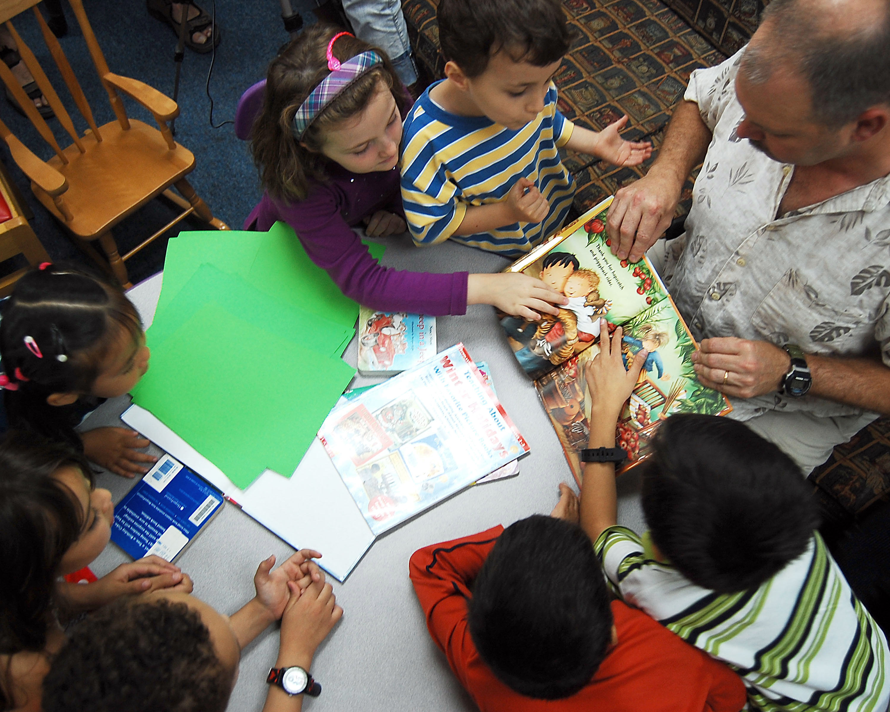 An adult reads a picture book aloud to young children around a library table.