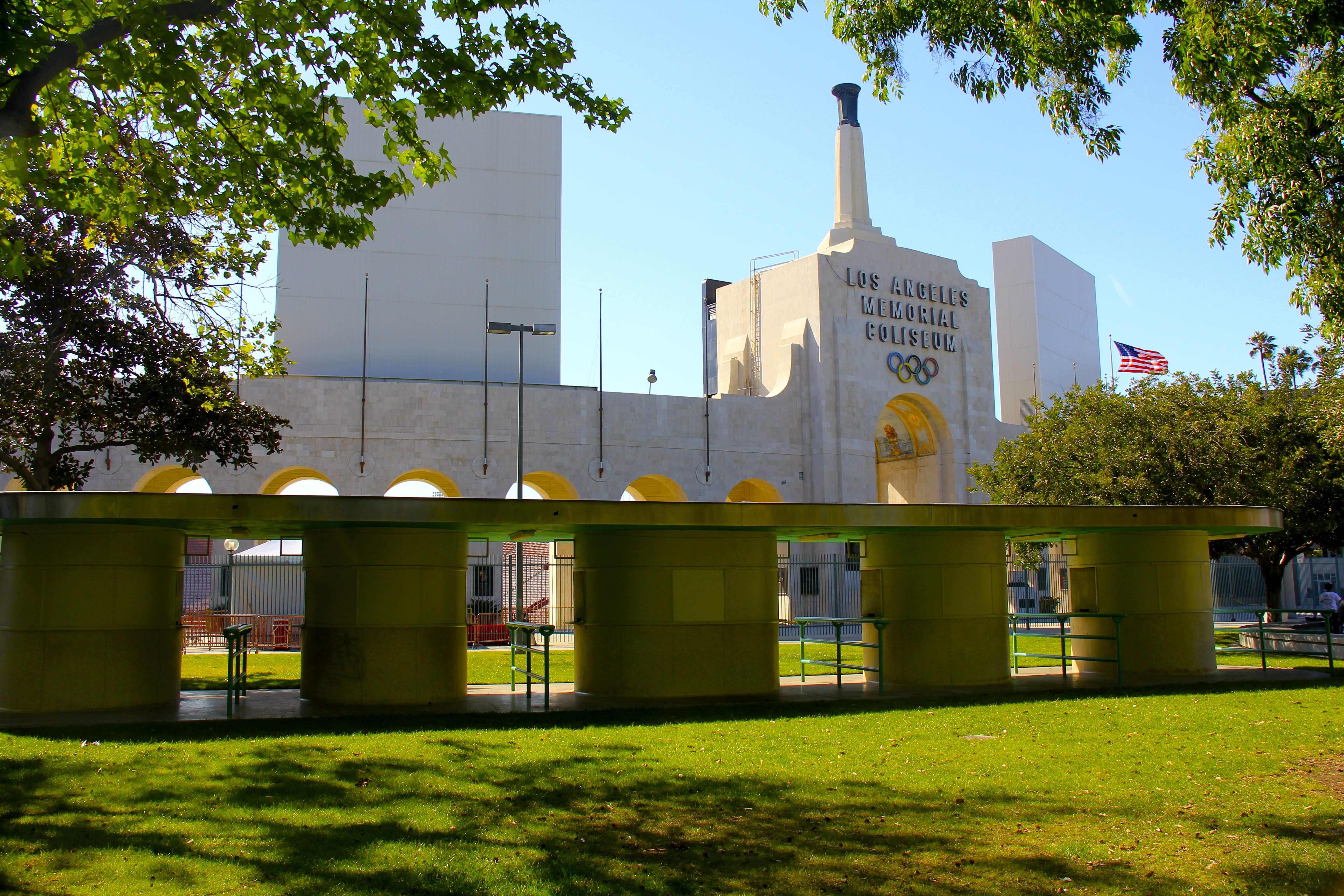 Exterior of a large stadium with Olympic rings and arched facade, framed by trees.